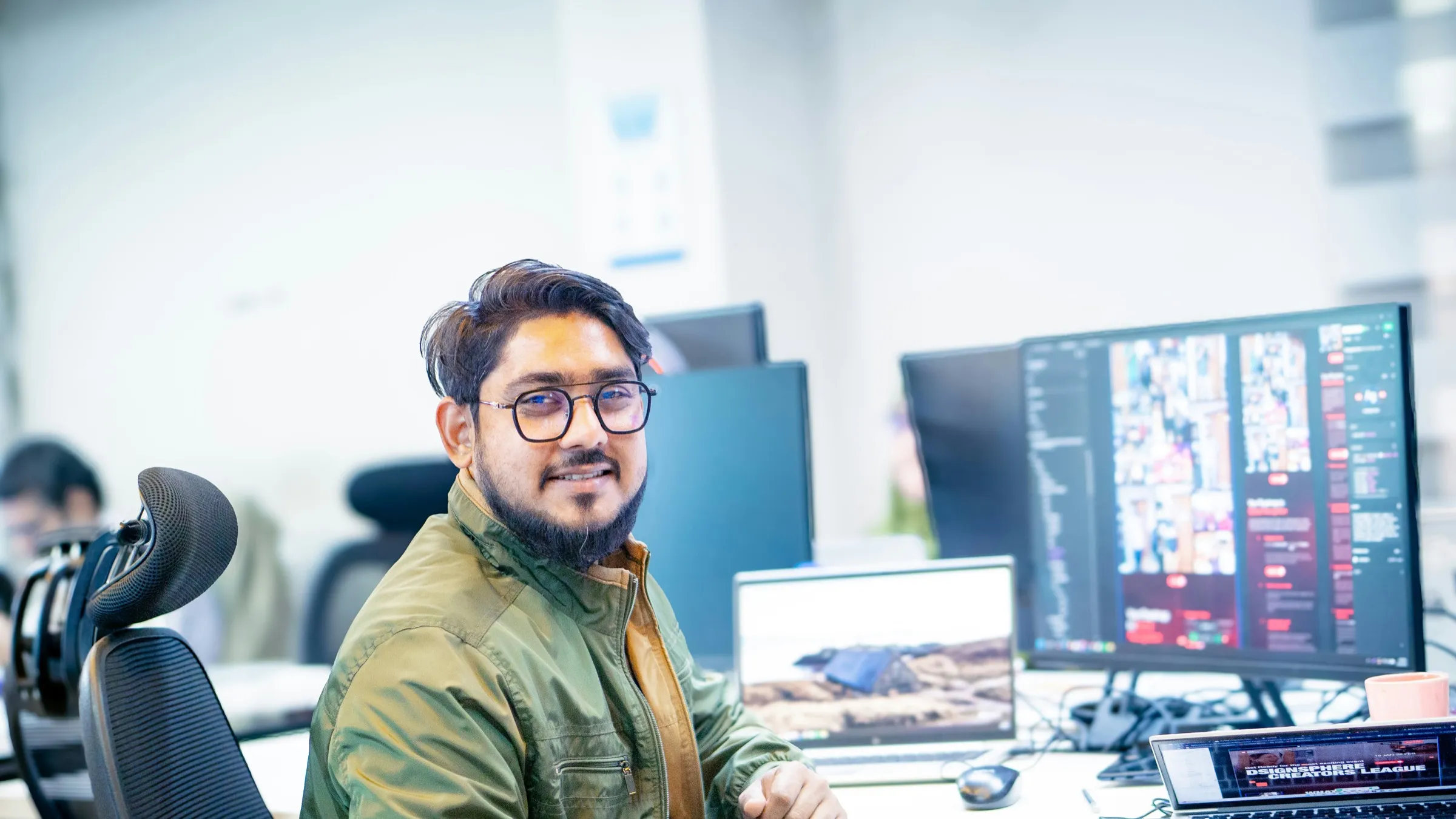 A developer at his desk in a busy office, monitors showing code